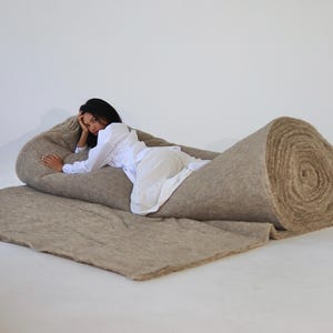 May include: A large, thick roll of natural beige fibrous material, partially unrolled to create a flat surface. The texture appears coarse and dense, with a light brown, earthy tone throughout the entire length of the material roll.