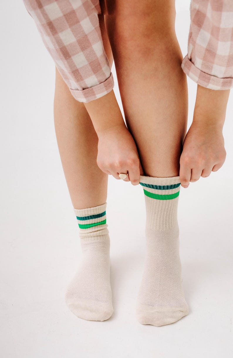 May include: Cream-colored socks with green and dark blue stripes at the top. The socks are being pulled up by a person wearing a pink and white checkered shirt. The socks are made of a soft, breathable material.