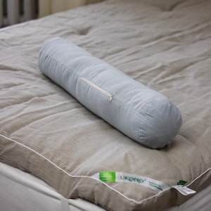 May include: A light blue cylindrical pillow with a zip closure rests on a beige mattress topper. The mattress topper is on a white mattress. The pillow and topper appear to be made of linen. The brand name "LIKONO" is visible on a tag.