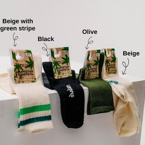 May include: Four pairs of socks in beige, black, and olive green. One pair is beige with a green stripe. Each pair has a tag with text in a foreign language and a cannabis leaf design. The socks are displayed on a white surface.
