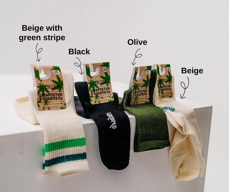 May include: Four pairs of socks in beige, black, and olive green. One pair is beige with a green stripe. Each pair has a tag with text in a foreign language and a cannabis leaf design. The socks are displayed on a white surface.