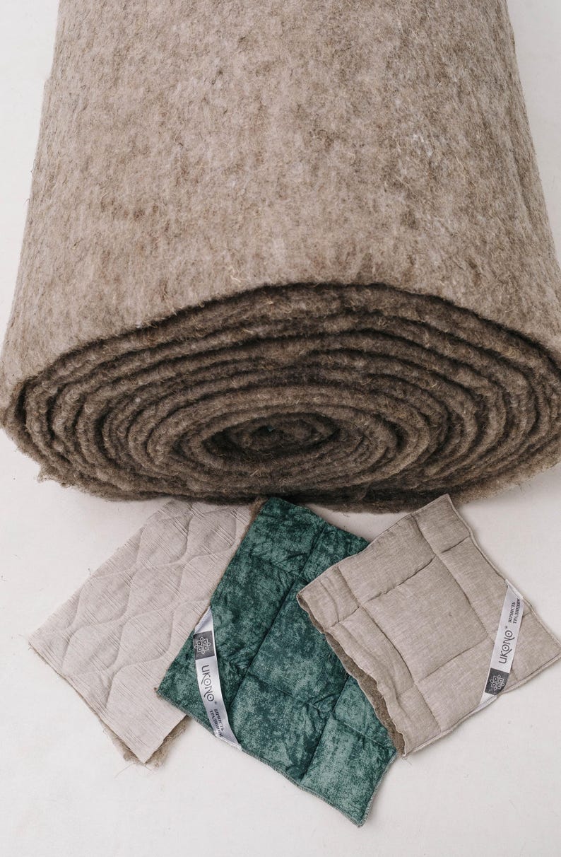 K&ouml;nnte beinhalten: Eine gro&szlig;e Rolle aus braunem Filzmaterial liegt &uuml;ber drei quadratischen, gesteppten Kissen in Beige und Gr&uuml;n. Die Kissen haben ein Rautenmuster und ein Etikett mit der Aufschrift "UKONO". Die Filzrolle scheint eine Naturfaser zu sein.