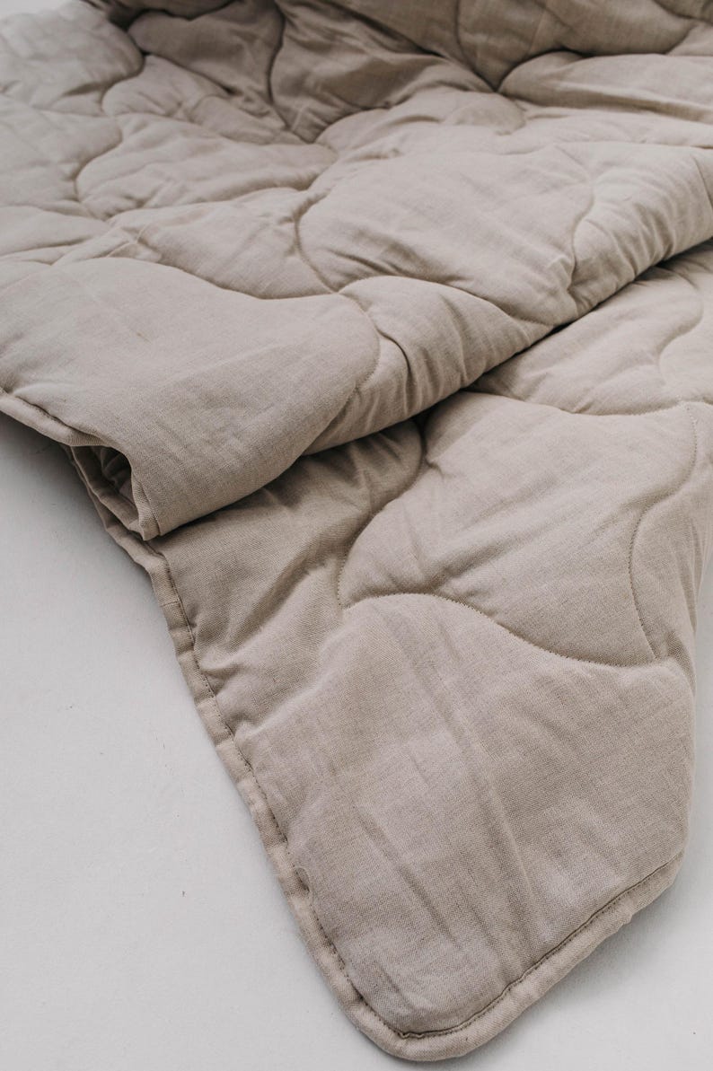 May include: A beige, quilted blanket with a soft, textured appearance. The blanket is folded over, showcasing the stitched pattern and the natural color of the fabric. The neutral tone suggests a focus on comfort and simplicity.