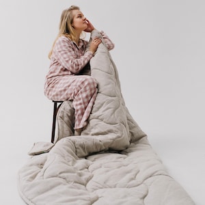 May include: A light beige quilted blanket with a matching pillow. The blanket is draped over a wooden stool. The person in the image is wearing a pink and white checkered pajama set.