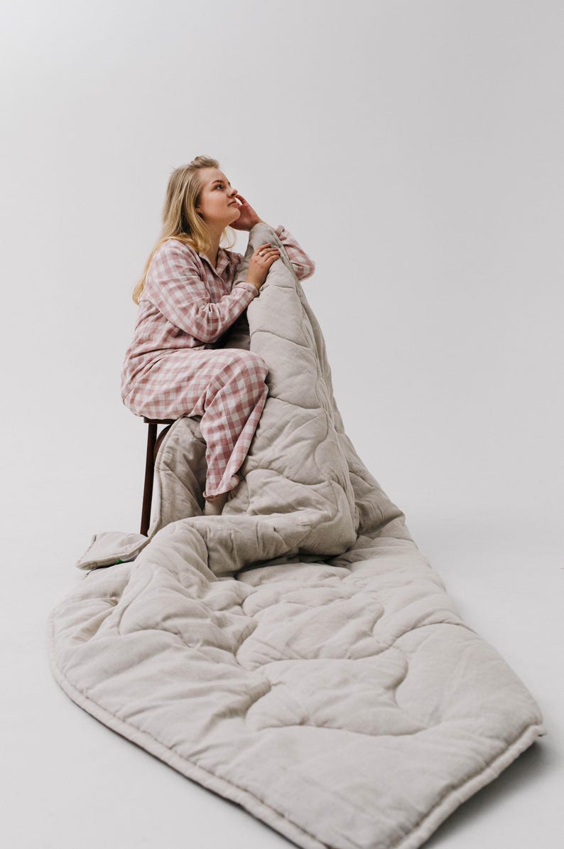 May include: A light beige quilted blanket with a matching pillow. The blanket is draped over a wooden stool. The person in the image is wearing a pink and white checkered pajama set.