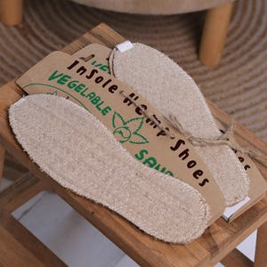Op de afbeelding: Een paar beige, gestructureerde stoffen inlegzolen bevestigd op een bruine kartonnen achterkant. De verpakking bevat groene en zwarte tekst met de tekst InSole Hemp Shoes en VEGETABLE, samen met een groene bladafbeelding en bruin touw.