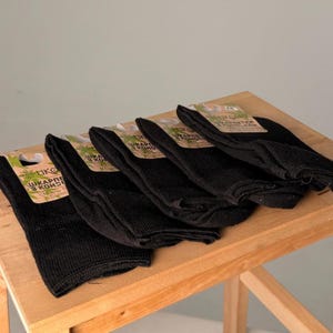 May include: A stack of five black socks with tags attached, displayed on a wooden surface. The tags have green and brown accents with text. The socks are folded and appear to be made of a thick, knitted material.