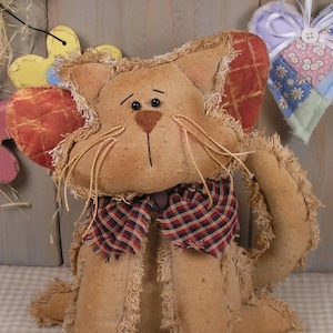 Könnte beinhalten: Eine handgemachte Stoffpuppe Katze mit einer rot-weiß karierten Schleife. Die Katze ist aus braunem Stoff mit einer zotteligen Textur und hat schwarze Knopfaugen. Die Katze sitzt auf einem karierten Stoffhintergrund. Das Wort "Pattern" ist in schwarzen Buchstaben am unteren Rand des Bildes geschrieben.