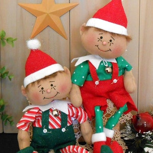 May include: Two handmade elf dolls with red and green outfits, white hats, and bells. The elves are sitting on a white crocheted doily.