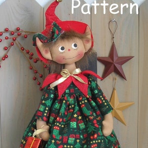 Puede incluir: Una muñeca de elfo de Navidad hecha a mano que lleva un vestido verde con estampado navideño rojo y blanco, un gorro rojo con una campana dorada y zapatos rojos con adornos dorados. La muñeca sostiene una pequeña caja de regalo roja. La muñeca está sentada en una silla de madera frente a una valla de madera.