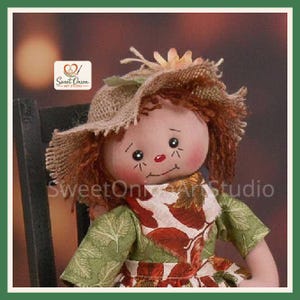 May include: A fabric doll with auburn yarn hair wearing a tan burlap hat and a green dress featuring a white apron with a brown and orange autumn leaf pattern. The doll has a painted face with black eyes, a red nose, and a stitched smile.