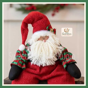 May include: A fabric Santa Claus doll wearing a red textured hat with a white pom-pom, a red textured body, and green and red plaid sleeves. The doll has a thick white yarn beard, black mittens, and a logo reading Sweet Onion Art Studio.