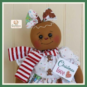 May include: A fabric gingerbread doll with a round brown face, black eyes, and rosy cheeks. It wears a white dress with colorful sprinkles, a red and white striped scarf, and a bow on its head. A tag attached reads Christmas love.