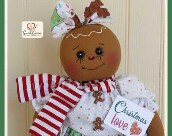 May include: A fabric gingerbread doll with a round brown face, black eyes, and rosy cheeks. It wears a white dress with colorful sprinkles, a red and white striped scarf, and a bow on its head. A tag attached reads Christmas love.