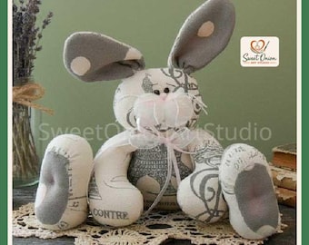 May include: A stuffed fabric rabbit toy featuring a grey and white pattern with script text and Eiffel Tower illustrations. The rabbit has long ears with light pink accents, a white ribbon bow at the neck, and white plush whiskers.