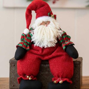 May include: A plush Santa Claus figurine wearing a red hat with a white pom-pom, a red and green plaid shirt, and red pants with black boots. The figure has a thick white yarn beard, a small nose, and gold jingle bells attached to the hat and pants.