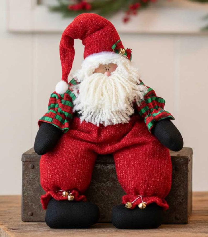 May include: A plush Santa Claus figurine wearing a red hat with a white pom-pom, a red and green plaid shirt, and red pants with black boots. The figure has a thick white yarn beard, a small nose, and gold jingle bells attached to the hat and pants.