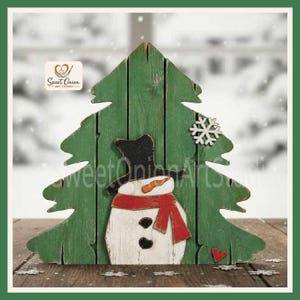 May include: A green wooden cutout shaped like a Christmas tree featuring a white snowman with a black top hat, an orange carrot nose, and a red scarf. A white snowflake and a small red heart are attached to the tree. The logo Sweet Onion Art Studio is visible.