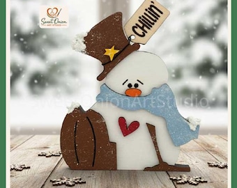 May include: A white wooden snowman decoration featuring a brown top hat with a yellow star, a light blue scarf, and a red heart on its chest. The snowman holds a brown pumpkin and has a wooden tag attached that reads "CHILLIN" in black text.