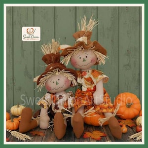 May include: Two fabric scarecrow dolls with straw hair and brown hats. The smaller doll wears a patterned shirt with sunflowers, while the larger doll wears a dress with an orange leaf print. Both dolls have stitched facial features.