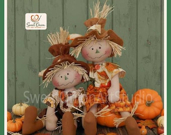 May include: Two fabric scarecrow dolls with straw hair and brown hats. The smaller doll wears a patterned shirt with sunflowers, while the larger doll wears a dress with an orange leaf print. Both dolls have stitched facial features.
