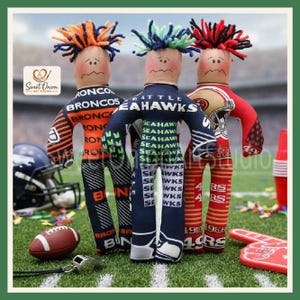 May include: Three fabric dolls with yarn hair, each wearing a different team-themed outfit. The left doll features orange and navy Broncos branding, the centre doll displays Seattle Seahawks text, and the right doll shows 49ers graphics.