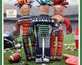May include: Three fabric dolls with yarn hair, each wearing a different team-themed outfit. The left doll features orange and navy Broncos branding, the center doll displays Seattle Seahawks text, and the right doll shows 49ers graphics.
