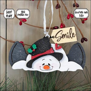 May include: A wooden snowman ornament with a black glitter top hat, red scarf, and a tag that says "Smile." The snowman has a white body with black and gray wings. The ornament is hanging from a white ribbon.