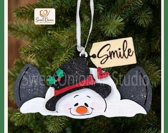 May include: A snowman ornament featuring a white face, a black glittery hat with a red band and green holly, and a small red heart. A wooden tag attached by a white ribbon displays the word Smile in black script. The ornament is flat.