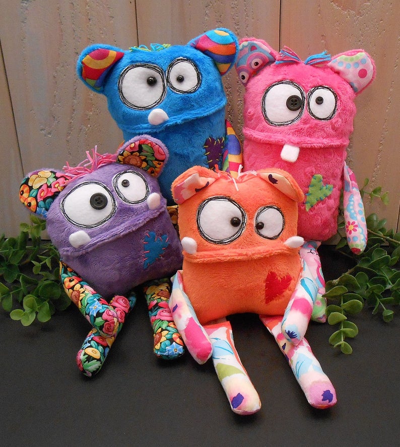 May include: Four plush monster toys in blue, pink, purple, and orange. Each creature features large white eyes with black button pupils, small white teeth, and long limbs made from various colourful floral and patterned fabrics with stitched patches.