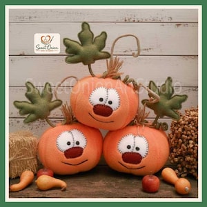 May include: Three plush orange pumpkins with stitched white circular eyes, dark brown noses, and embroidered smiles. Each pumpkin features a green felt leaf and a brown twine stem. The text Sweet Onion Art Studio is visible in the corner.