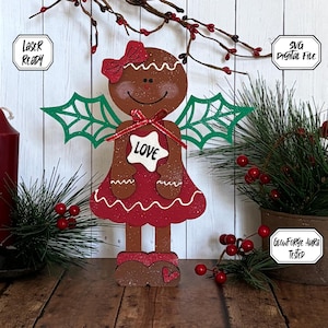 Puede incluir: Un ángel de jengibre con alas de acebo verdes, un vestido rojo y una estrella blanca con la palabra "LOVE" en ella. El ángel está de pie sobre una superficie de madera.