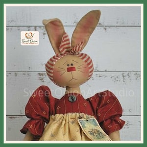 May include: A fabric rabbit doll with long ears, wearing a red and white striped headpiece, a dark red puff-sleeved dress with star patterns, and a yellow skirt. A small tag is attached to the dress, and a Sweet Onion Art Studio logo is visible.