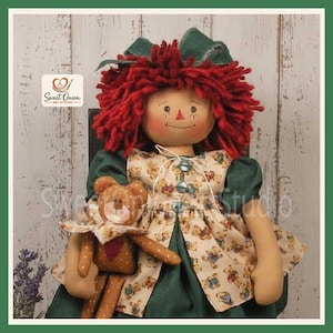 May include: A fabric doll with bright red yarn hair and a dark green bow, wearing a dark green dress with a beige apron featuring a small teddy bear pattern. The doll holds a small brown plush teddy bear. A logo reads Sweet Onion Art Studio.