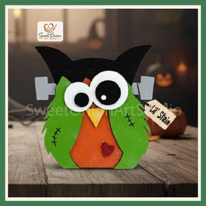 May include: A wooden owl decoration styled as a monster with a black head, green wings featuring black stitched details, and an orange body with a small red heart. It has large white eyes, grey bolts on the sides, and a tag reading Lil' Stein.