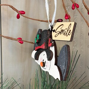 May include: A wooden snowman ornament with a black hat, red scarf, and orange nose. The ornament has a tag that says "Smile" and is hanging from a white ribbon. The background includes branches with red berries and green pine needles.