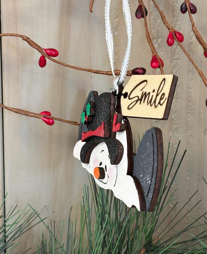 May include: A wooden snowman ornament with a black hat, red scarf, and orange nose. The ornament has a tag that says "Smile" and is hanging from a white ribbon. The background includes branches with red berries and green pine needles.