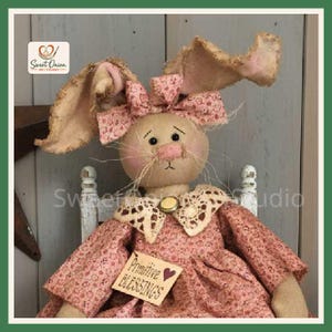 May include: A fabric rabbit doll with long, frayed ears, wearing a pink floral dress and a matching bow. The doll features a beige face with pink cheeks, a lace collar with a small button, and a tag that reads Primitive Blessings.