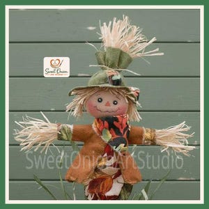 May include: A fabric scarecrow doll featuring a green felt hat with raffia hair, a brown jacket, and a scarf patterned with autumn leaves. The doll has a round face with stitched features and raffia hands, with the Sweet Onion Art Studio logo.