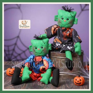 May include: Two green fabric dolls with stitched facial features and black hair. One doll sits on a metal container wearing a black shirt with white patterns, while the other sits in front wearing a blue shirt with orange pumpkin prints.