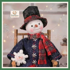 Puede incluir: Una figura de muñeco de nieve de tela con un sombrero de copa negro brillante con una banda de cuadros rojos, un abrigo azul oscuro con botones de copo de nieve y una bufanda de cuadros rojos. Sostiene un adorno de copo de nieve de felpa.