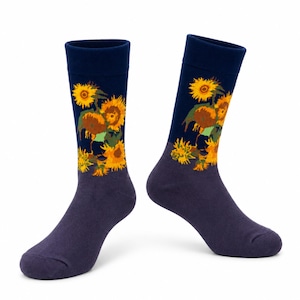 Op de afbeelding: Een paar kuitlange sokken met een donkerblauw bovengedeelte voorzien van een levendige gele en oranje zonnebloemenprint. Het onderste gedeelte, inclusief de hiel en de teen, is effen gedempt paars van kleur met een geribbelde boord aan de bovenkant.
