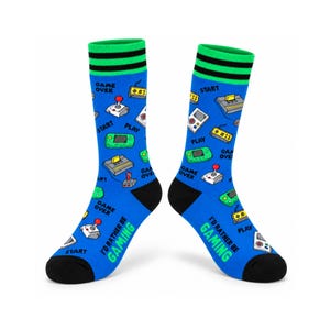 Puede incluir: Un par de calcetines azules con un patrón repetido de consolas de videojuegos retro, joysticks y dispositivos portátiles. El texto "I'D RATHER BE GAMING" está impreso en verde cerca de la puntera negra. Los puños tienen rayas verdes y negras.
