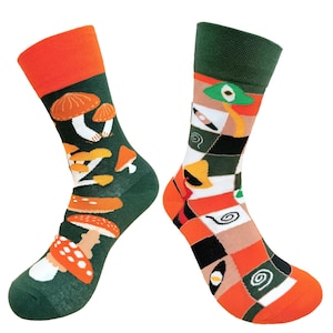 Op de afbeelding: Een paar novelty sokken met contrasterende oranje en groene manchetten en tenen. Eén sok heeft een patroon van oranje en witte paddenstoelen op een groene achtergrond. De andere sok heeft een patchwork ontwerp met sushi en andere etenswaren.