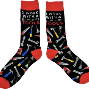 Könnte beinhalten: Ein Paar schwarze Socken mit rotem Bund. Die Socken haben ein weißes und rotes Design mit verschiedenen Werkzeugen, darunter Hämmer, Sägen, Schraubenschlüssel und Schraubendreher. Die Socken haben den Text "Ich arbeite mit einem Haufen Werkzeugen" aufgedruckt.