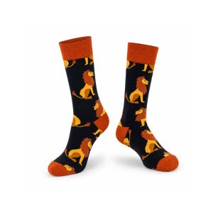 Peut inclure: Une paire de chaussettes mi-mollet avec une base noire et un motif répétitif de lions jaunes et marron. Les chaussettes présentent des bords-côtes, des talons et des pointes de couleur orange uni, contrastant avec le tissu de fond sombre.