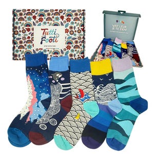 Könnte beinhalten: Eine Auswahl an bunten Socken mit verschiedenen Mustern, darunter Wellen, Berge und abstrakte Designs. Die Socken werden zusammen mit einer dekorativen Box mit der Aufschrift "Tutti Footi" und "Step Into Fun" präsentiert.