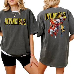 Op de afbeelding: Donkergrijs T-shirt met het woord "INVINCIBLE" in gele blokletters op de voorkant. De achterkant van het shirt toont een groep striphelden. Het T-shirt heeft een relaxte pasvorm en korte mouwen.