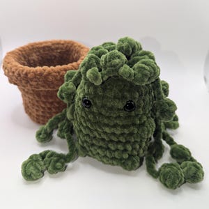 Puede incluir: Una criatura de peluche verde hecha a mano con ojos negros y zarcillos, junto a una maceta de peluche marrón. El juguete de peluche tiene un aspecto texturizado y difuso. La maceta es de color marrón claro y tiene una textura similar.
