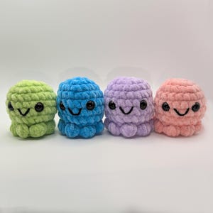 Puede incluir: Cuatro pequeños juguetes de pulpo de ganchillo. Cada pulpo es de un color diferente: verde, azul, morado y rosa. Tienen ojos negros y caras sonrientes. Los juguetes están hechos de un hilo suave y texturizado.